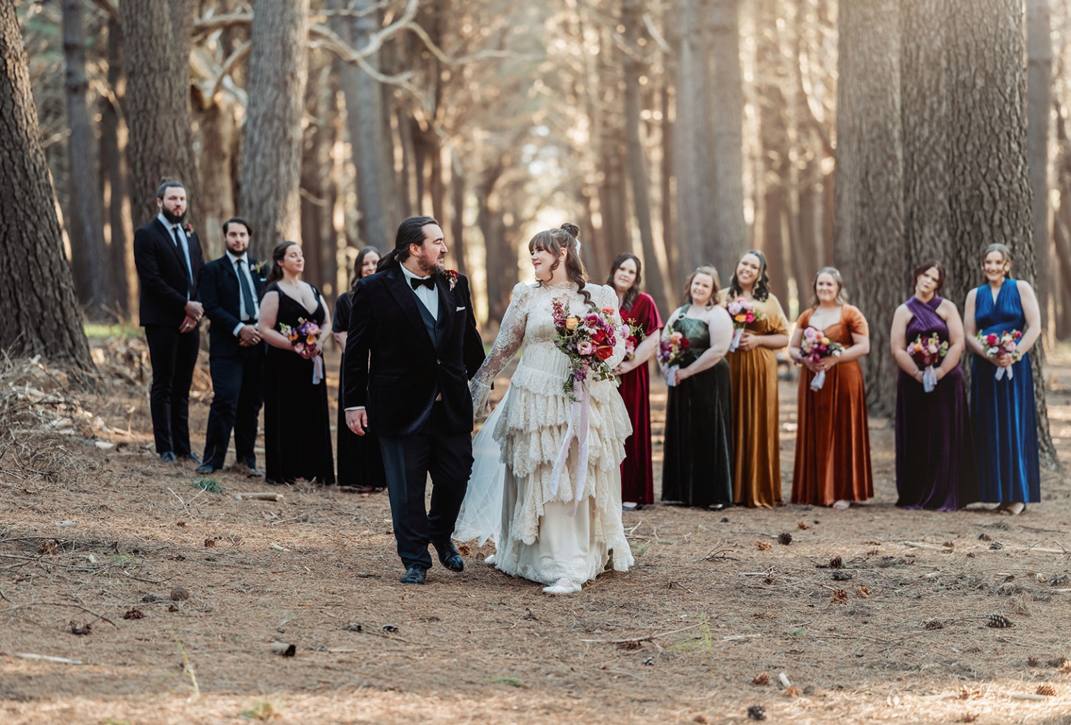 burbrook wedding – amber & angus | kuitpo forest wedding 37 Amber and Angus walk hand in hand in a forest, with their wedding party standing behind them holding colourful bouquets.