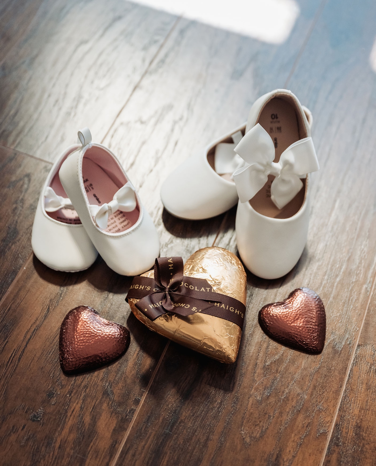 serafino winery wedding – maor & sasha 13 Two pairs of white baby shoes with bow details beside heart-shaped chocolates on a wooden floor.