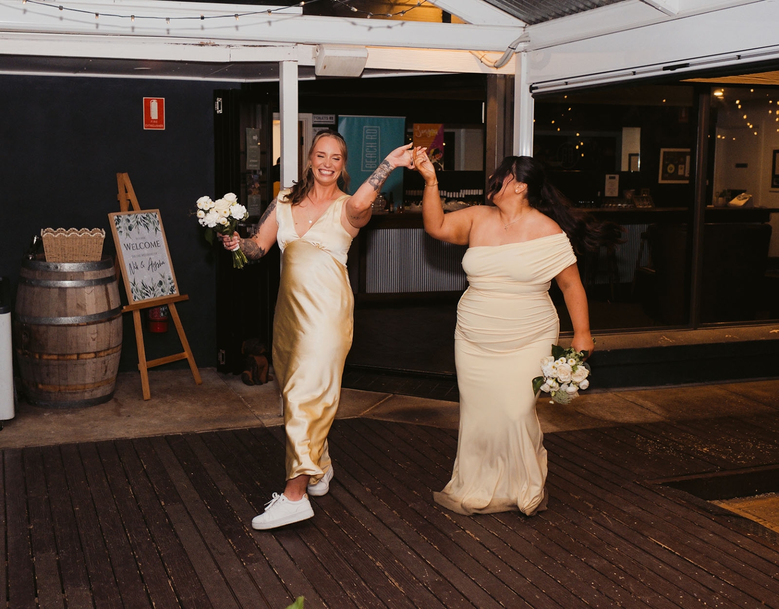 beach road winery wedding – alysha & nik 48 Two women in ivory dresses holding bouquets and dancing beneath festoon lights at Beach Road Winery
