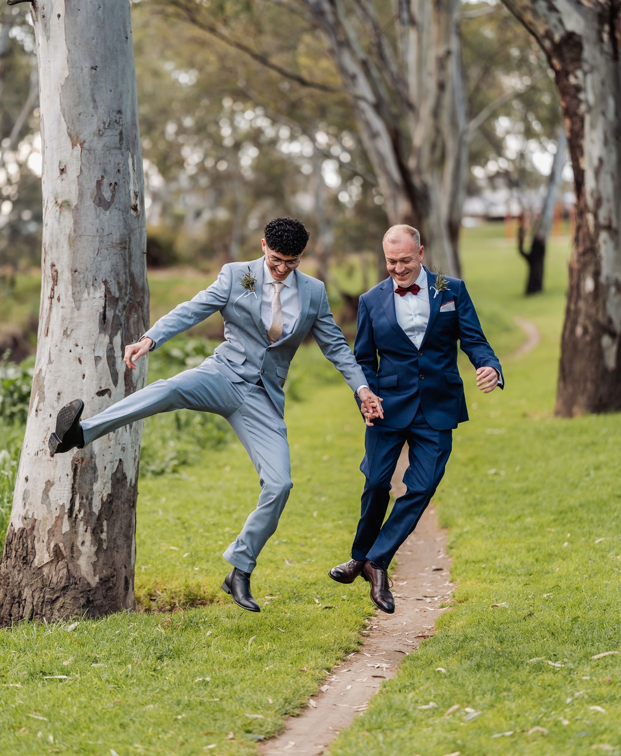 hotel royal wedding - brad & manuel 12 Brad and Manuel in wedding suits holding hands and jumping along a tree-lined park path