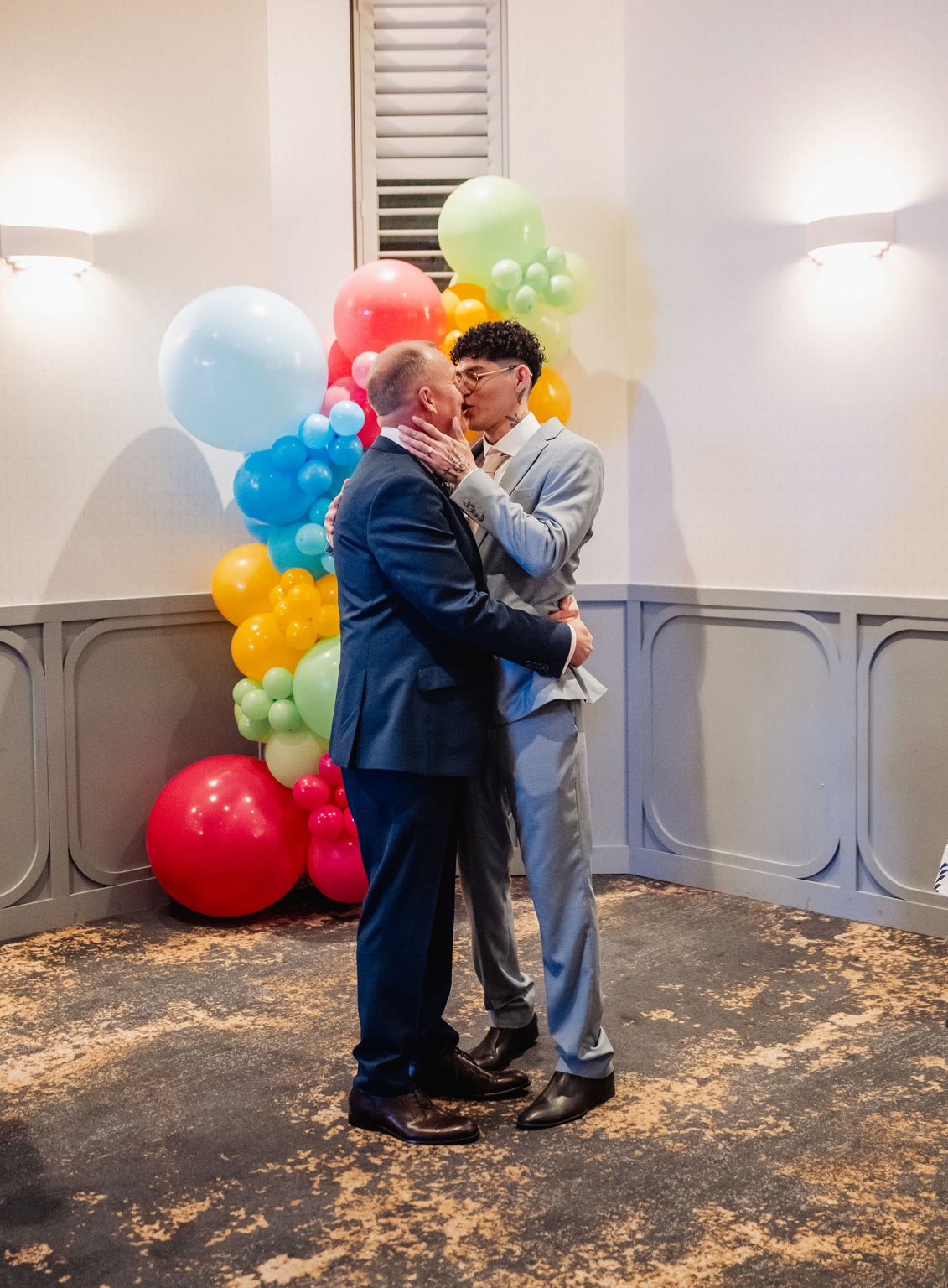 hotel royal wedding - brad & manuel 29 Brad and Manuel kissing in suits beside a colourful balloon display at Hotel Royal