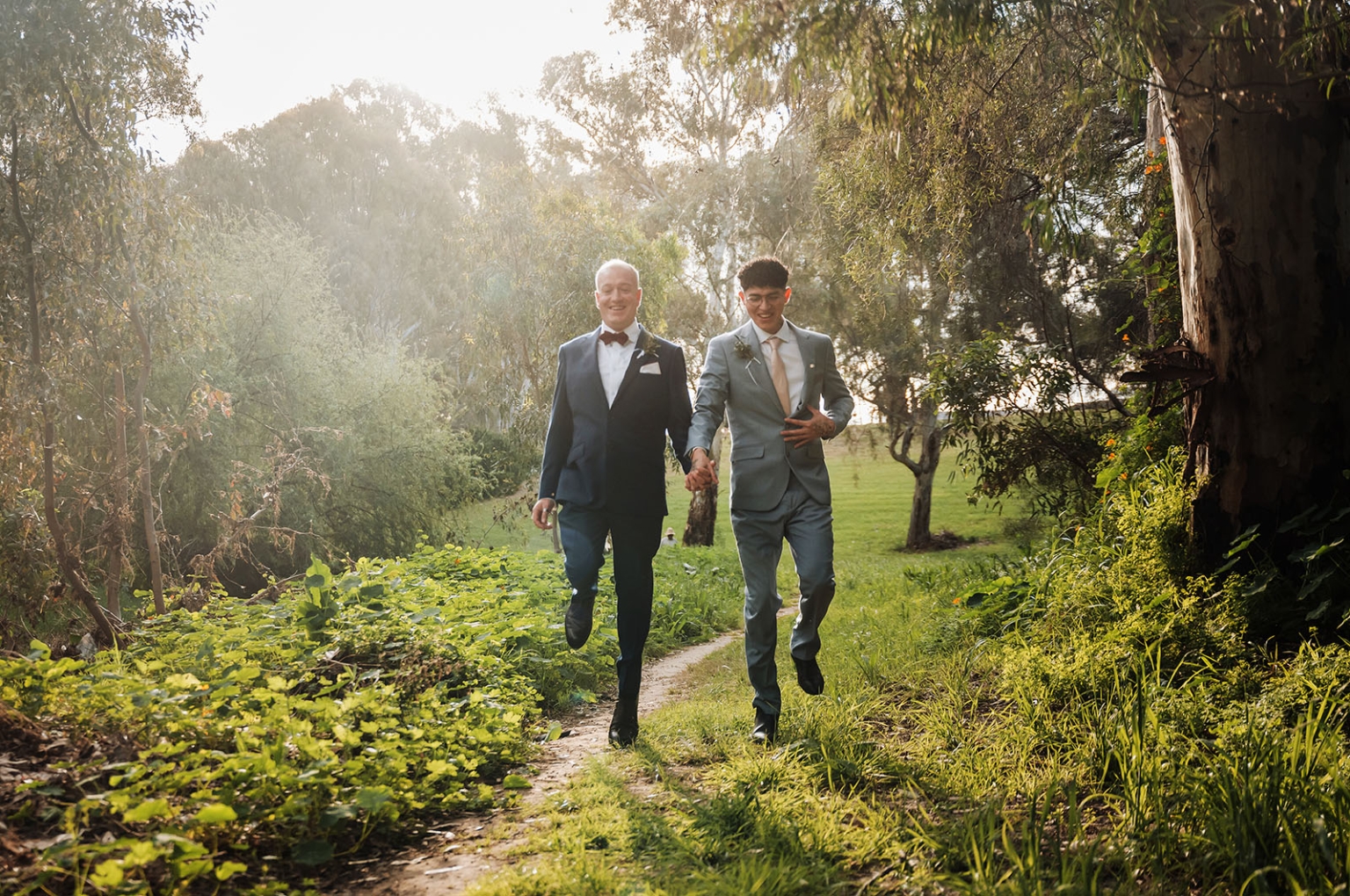 hotel royal wedding - brad & manuel 21 Brad and Manuel in suits walking hand in hand along a sunlit path through leafy parkland