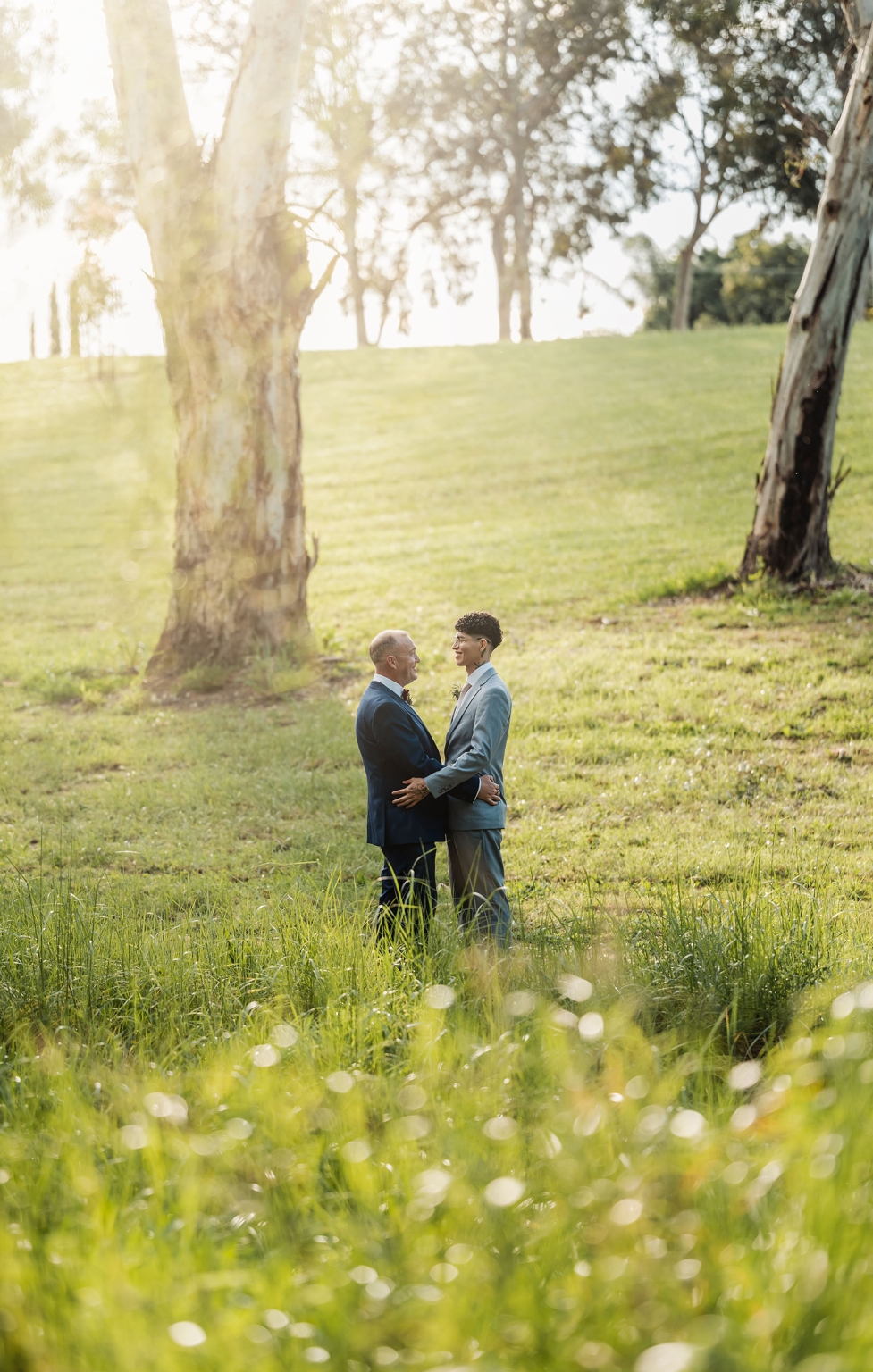 hotel royal wedding - brad & manuel 19 Brad and Manuel embracing in suits beneath gum trees in a sunlit grassy park.