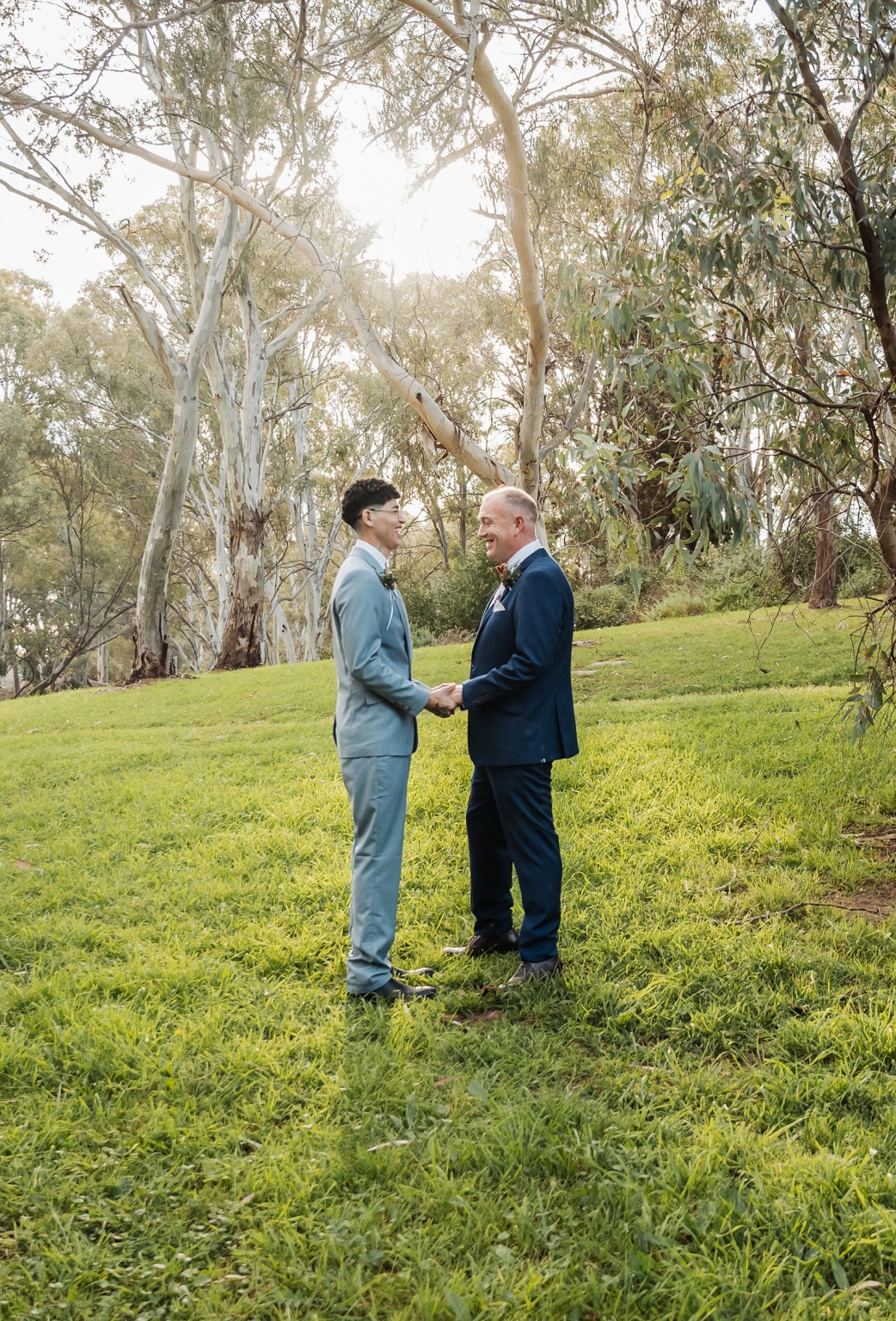 hotel royal wedding - brad & manuel 13 Brad and Manuel holding hands and smiling in suits beneath sunlit gum trees in a park