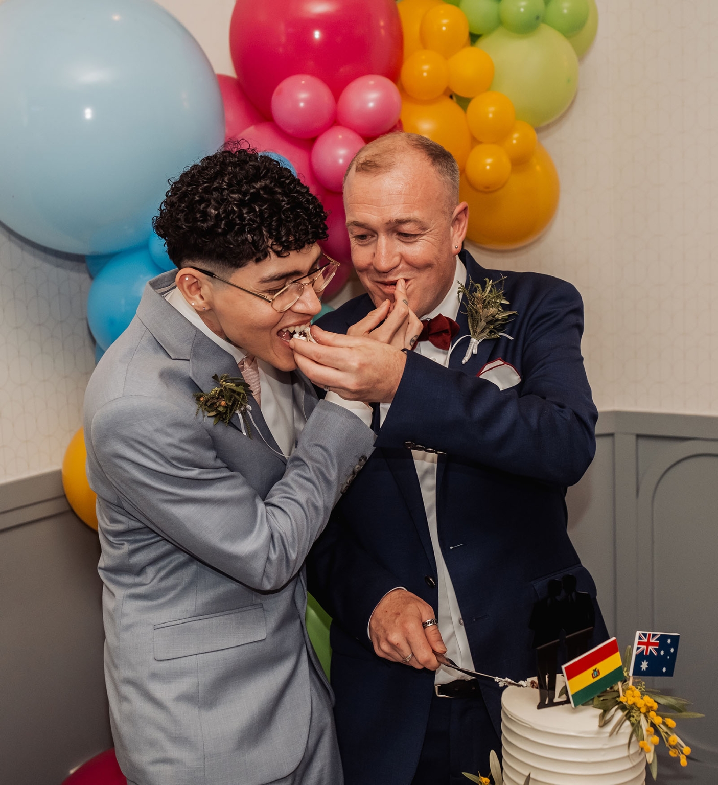 hotel royal wedding - brad & manuel 41 Brad and Manuel in suits feeding each other wedding cake beside colourful balloons