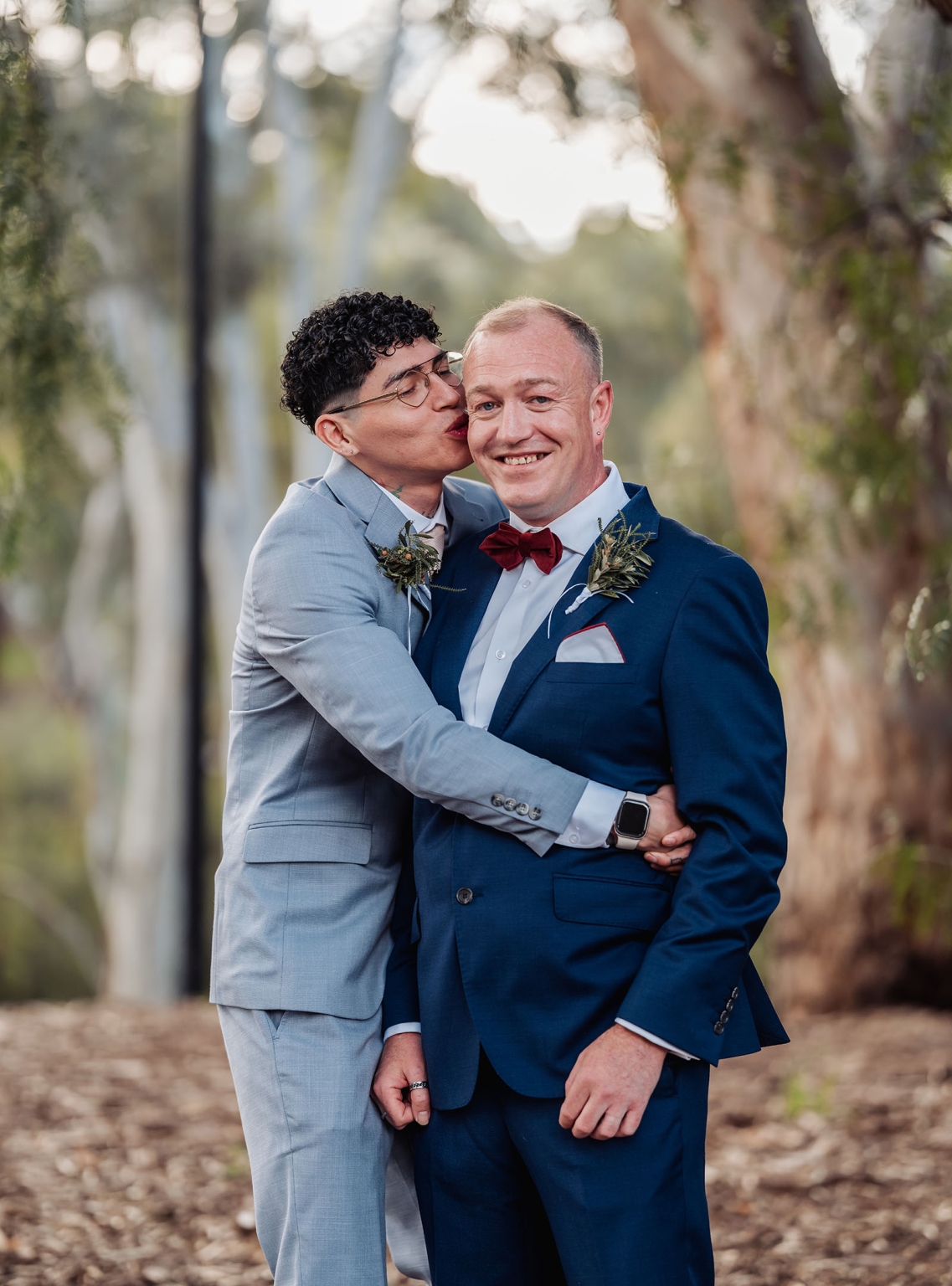hotel royal wedding - brad & manuel 11 Brad kisses Manuel on the cheek as they embrace in suits among trees during their wedding portrait session.
