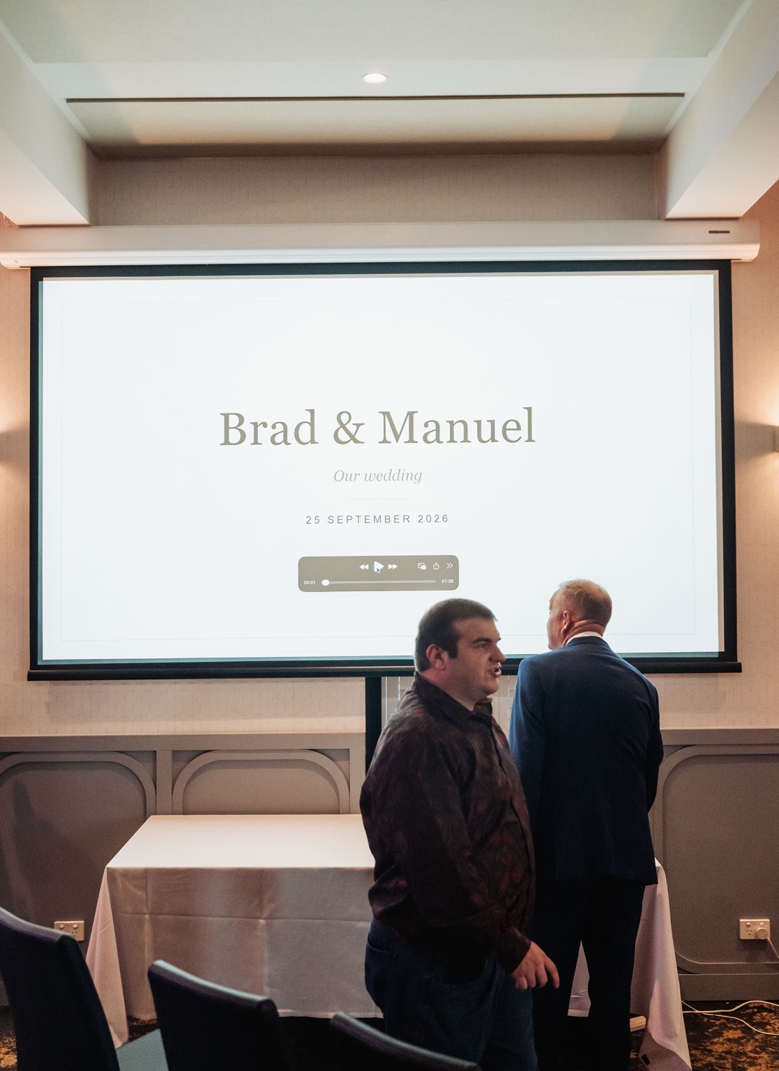 hotel royal wedding - brad & manuel 22 Two guests stand before a projection screen displaying Brad and Manuel's wedding slideshow and date.
