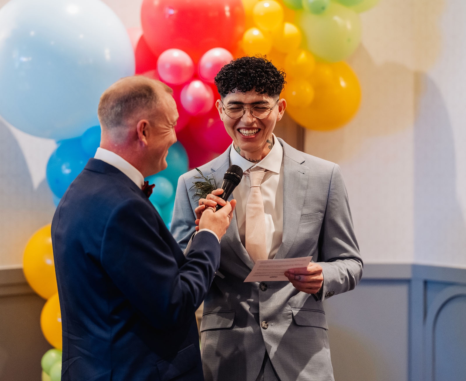 hotel royal wedding - brad & manuel 26 Two grooms laughing during their wedding vows, one holding a microphone and the other reading from a card before colourful balloons.