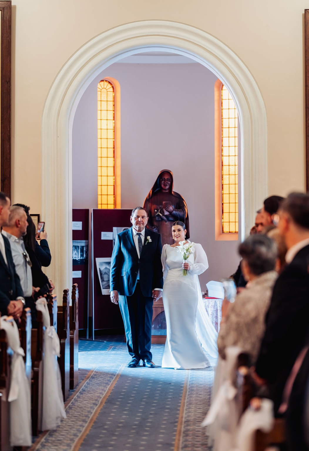 st joseph's chapel wedding – diana & michael 9 Bride in a white gown holding a lily bouquet walks down the aisle with a man in a dark suit at St Joseph's Chapel