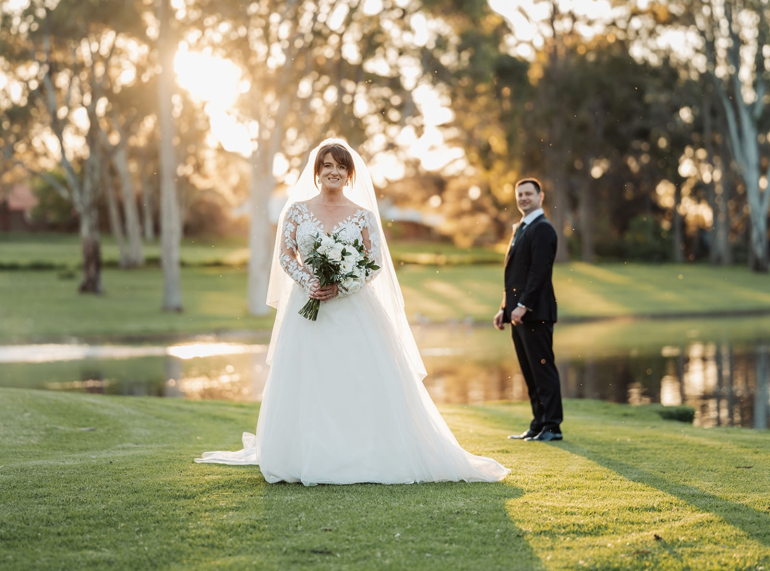 serafino winery wedding – maor & sasha 50 Bride holding a white bouquet on a lawn beside a pond at sunset, with the groom standing behind her