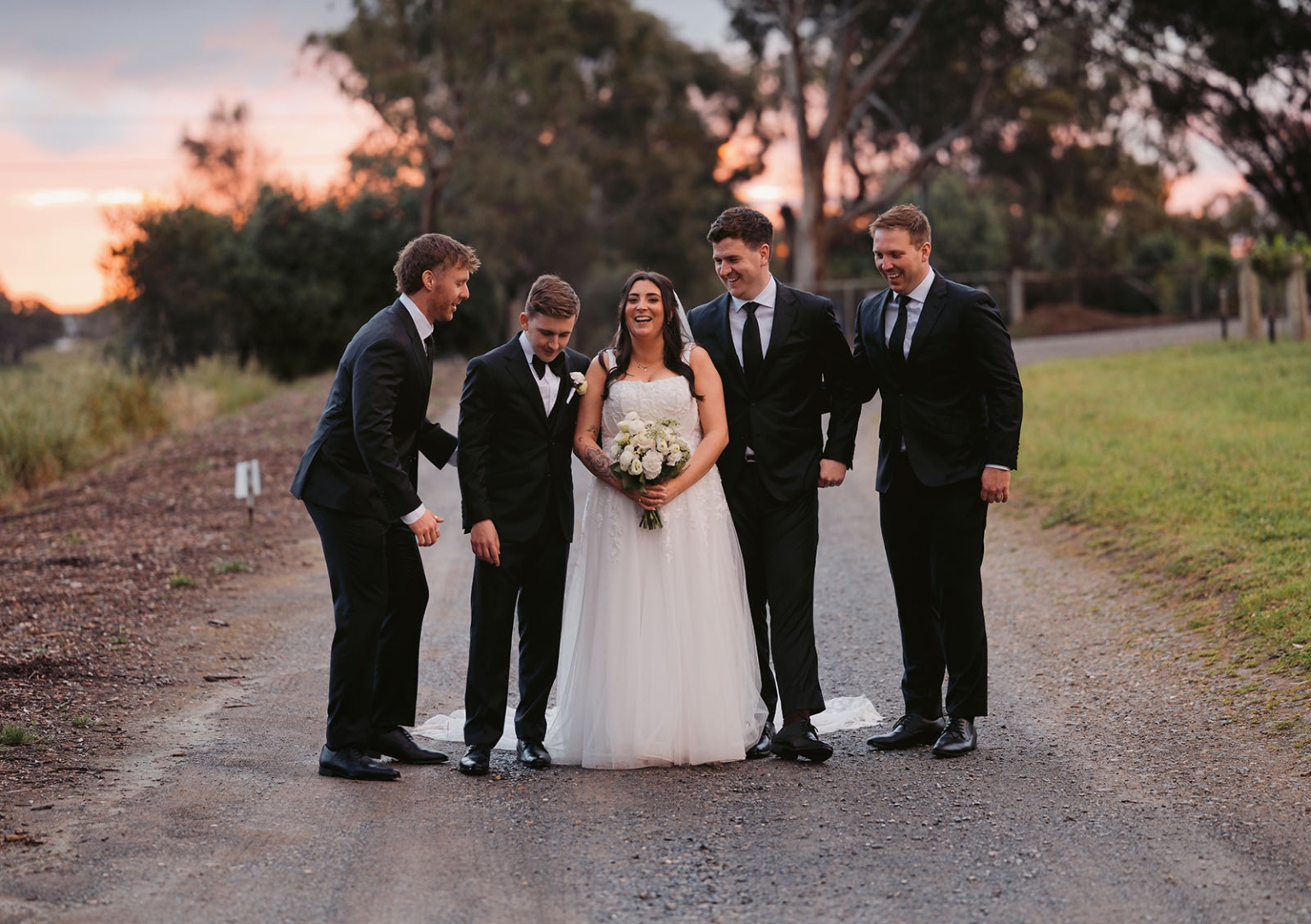 beach road winery wedding – alysha & nik 37 Bride holding a white bouquet laughs with four groomsmen in black suits on a country lane at sunset at Beach Road Winery.