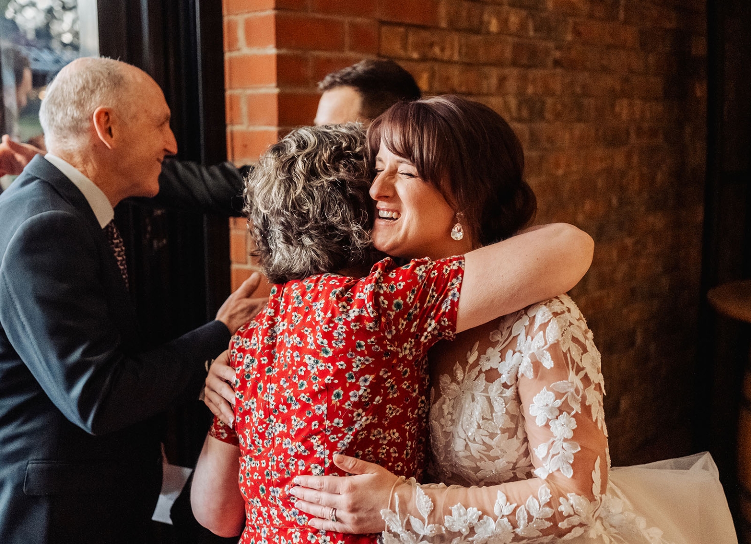 serafino winery wedding – maor & sasha 58 Bride in a lace wedding gown smiles while hugging a guest in a red floral dress indoors.