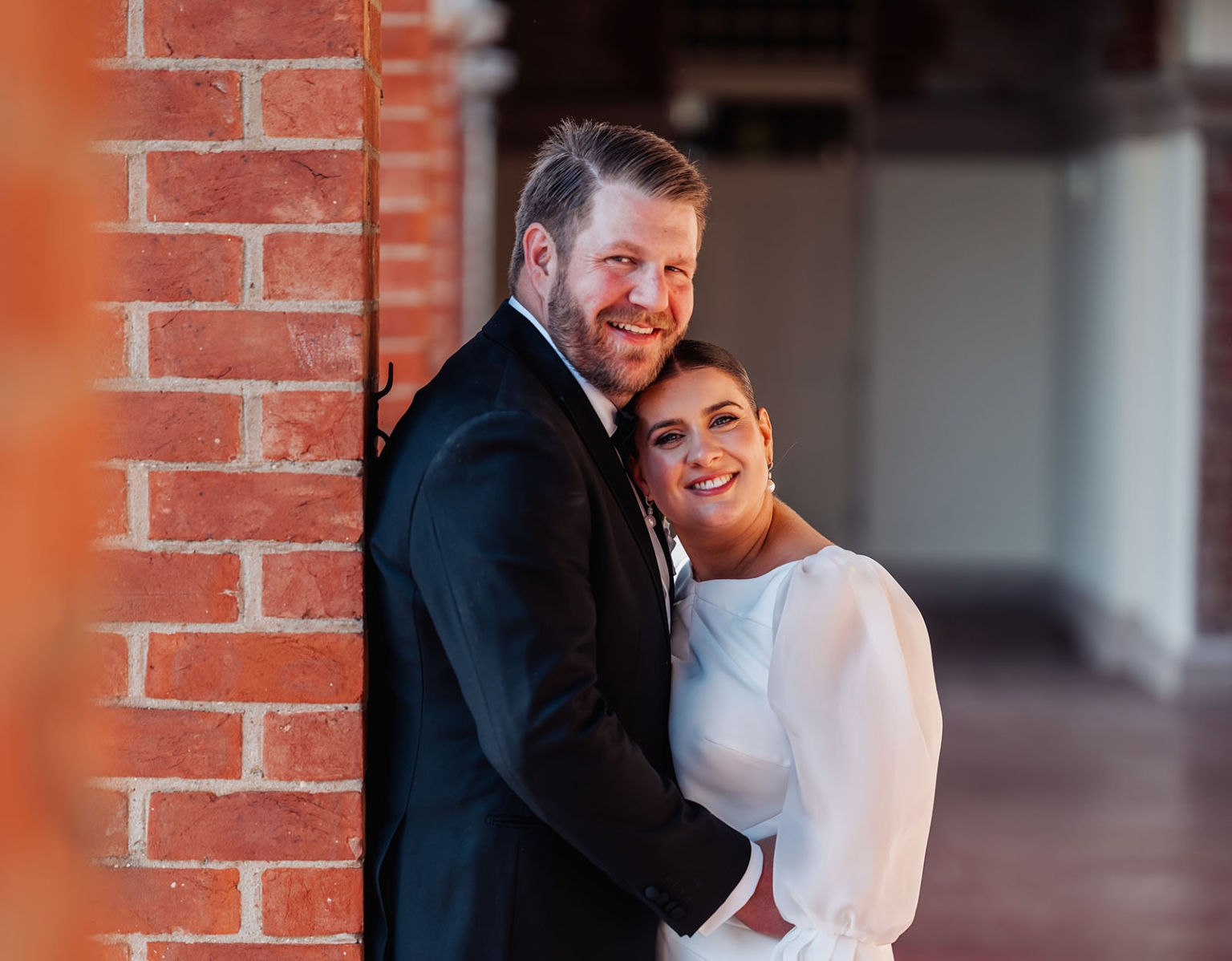 st joseph's chapel wedding – diana & michael 25 Diana and Michael smiling together beside a red brick wall, with Michael in a black suit and Diana in a white wedding dress.