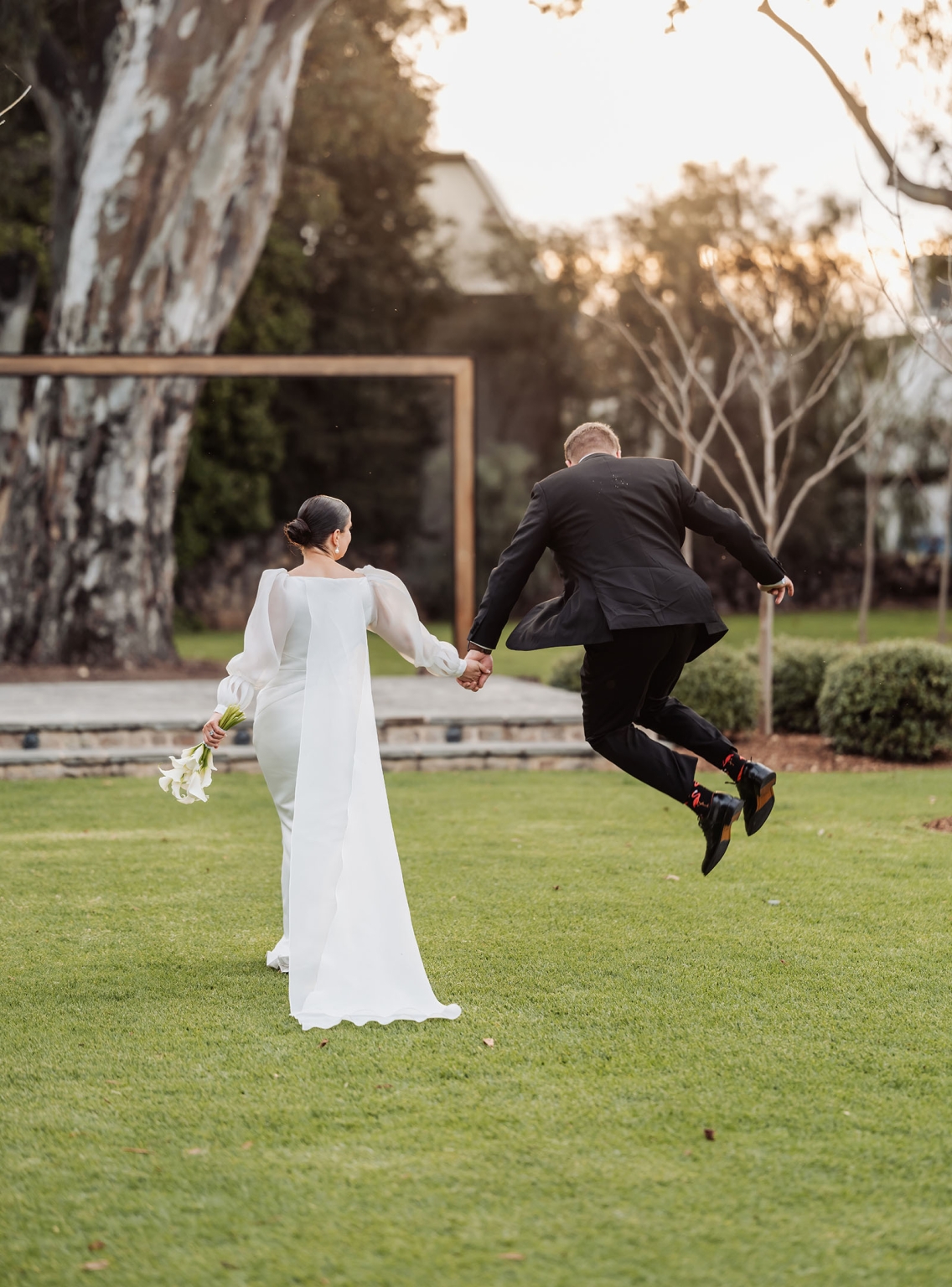 st joseph's chapel wedding – diana & michael 37 Bride in a long white gown holds hands with the groom as he jumps across a lawn at their garden wedding.
