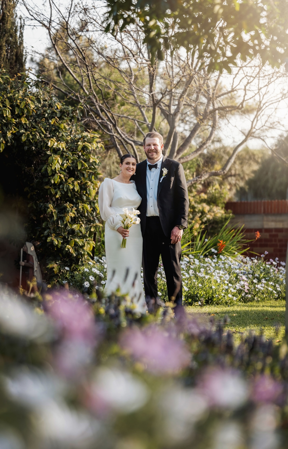 st joseph's chapel wedding – diana & michael 30 Diana and Michael standing together in a sunlit garden, with Diana holding a white bouquet and Michael wearing a black tuxedo.