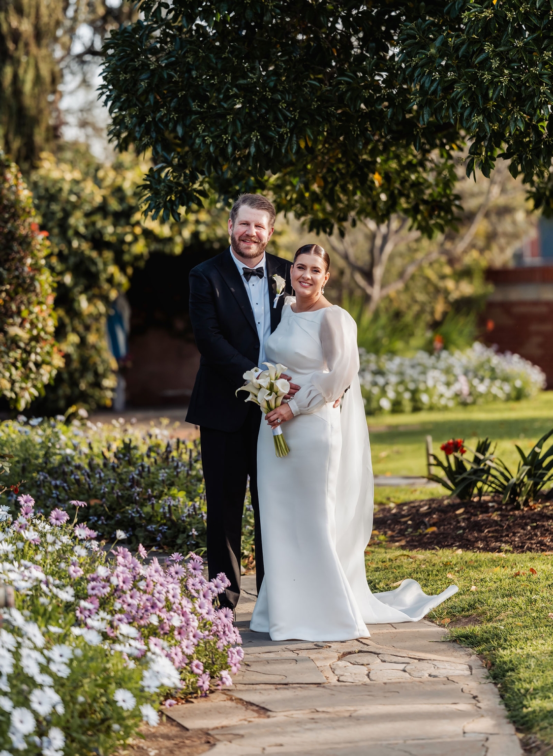 st joseph's chapel wedding – diana & michael 29 Diana and Michael standing together on a garden path beside pink and white flowers after their wedding ceremony.