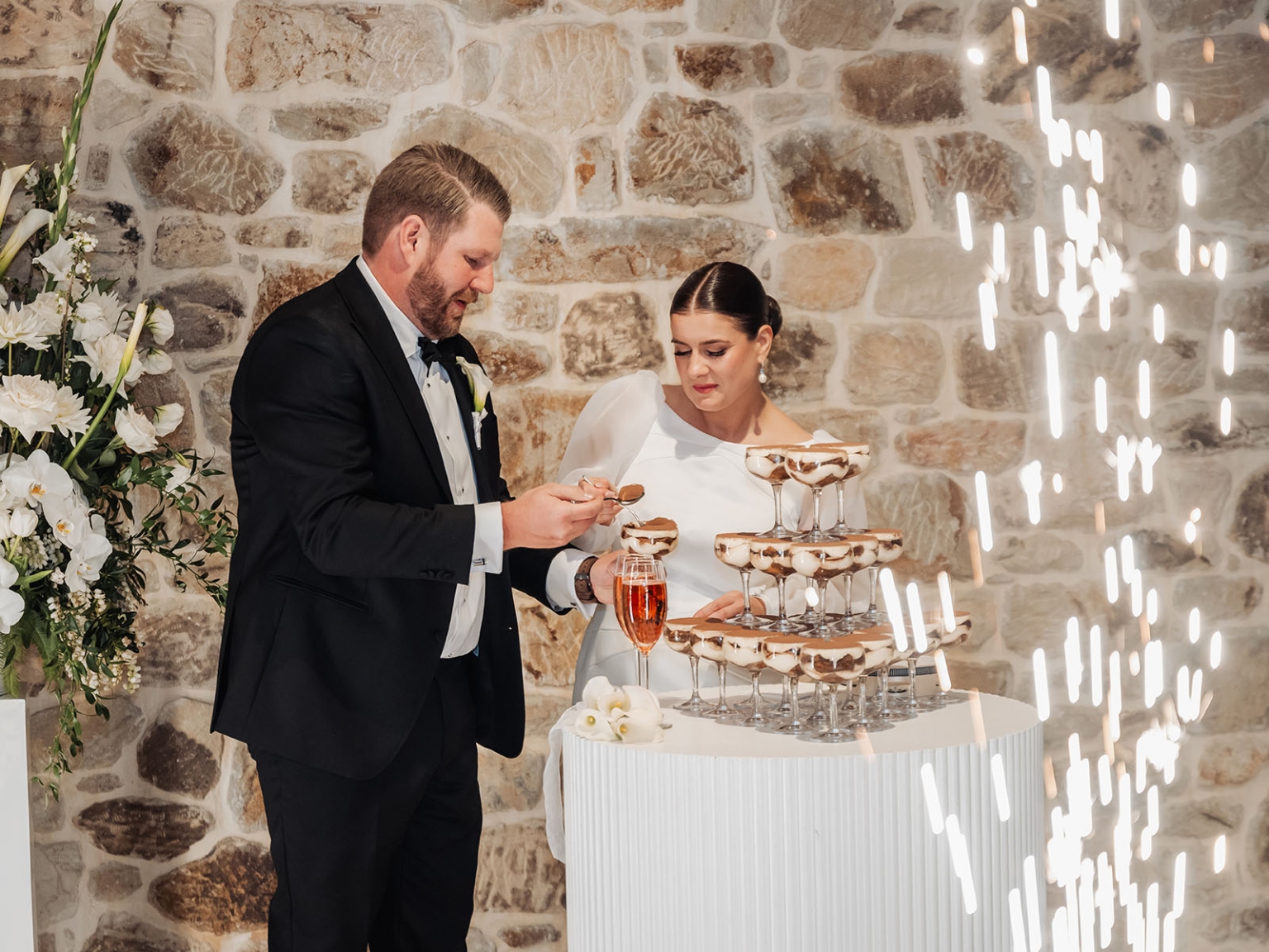 st joseph's chapel wedding – diana & michael 46 Diana and Michael serving layered desserts in coupe glasses beside a white floral arrangement at their wedding reception
