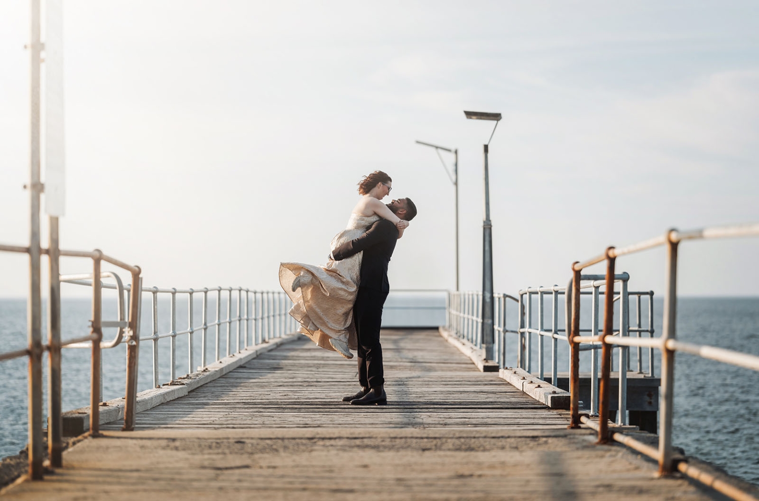 Punit lifts Ebony in her wedding dress on a wooden pier above the sea at Second Valley.