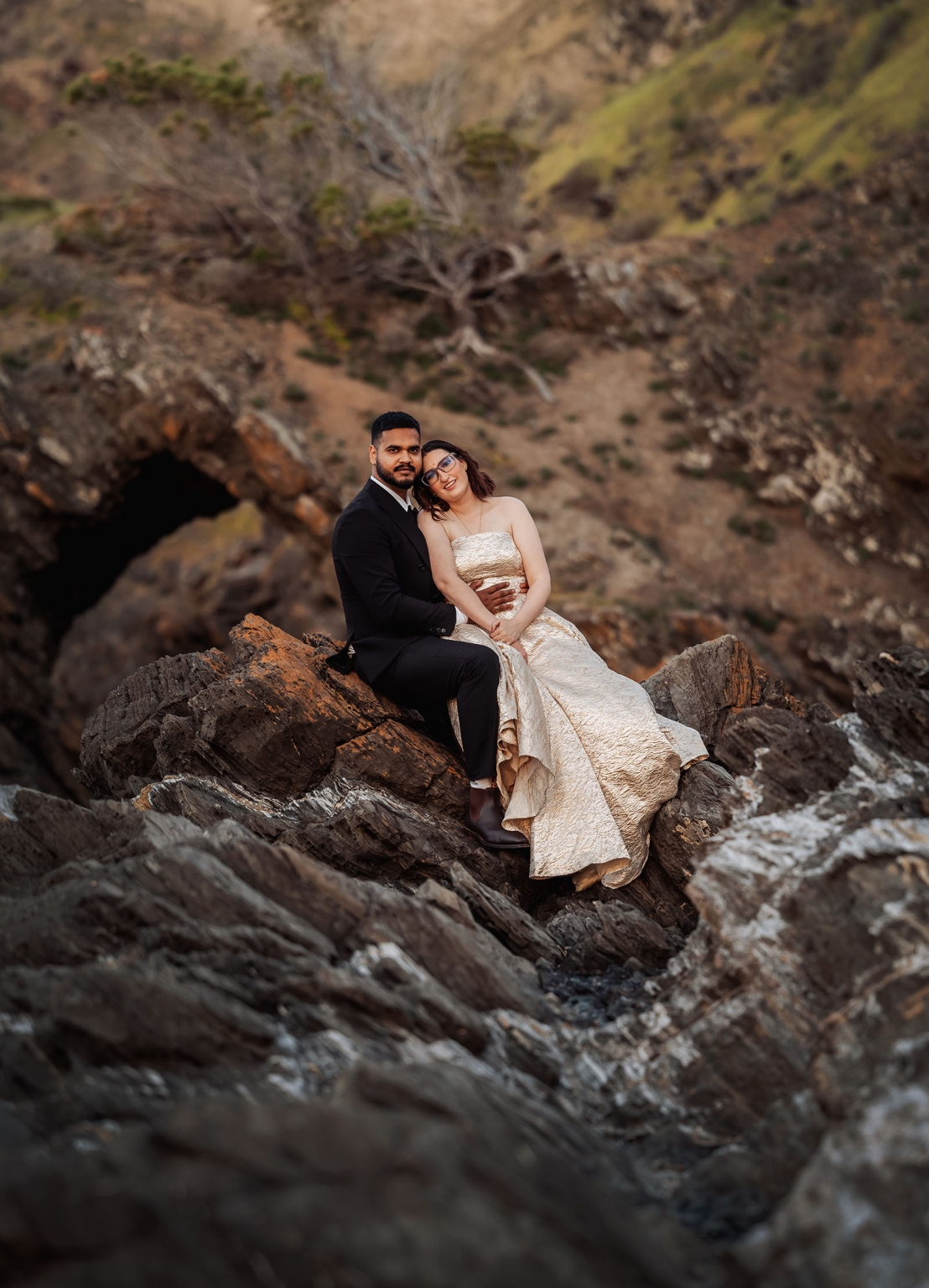 Ebony and Punit seated together on rugged coastal rocks, wearing a wedding dress and black suit at Second Valley.