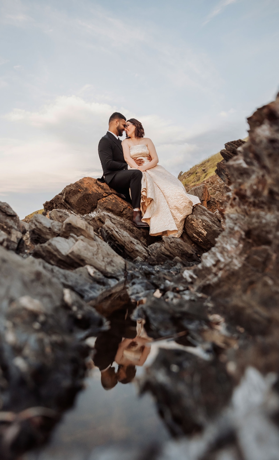 Ebony and Punit sitting close together on rocky ground in wedding attire, reflected in a small puddle at Second Valley