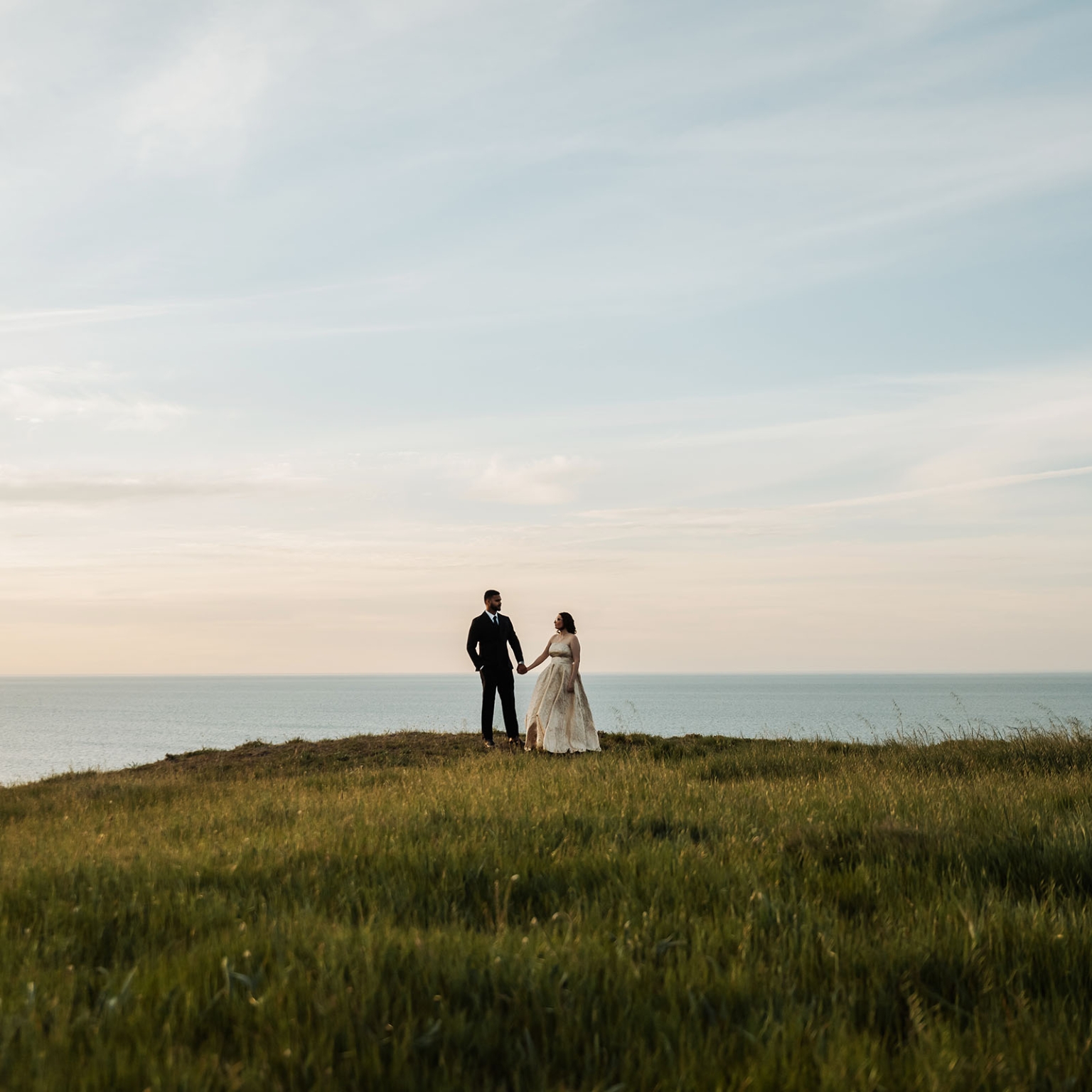 Ebony and Punit holding hands on a grassy clifftop above the sea, dressed in wedding attire at Second Valley.