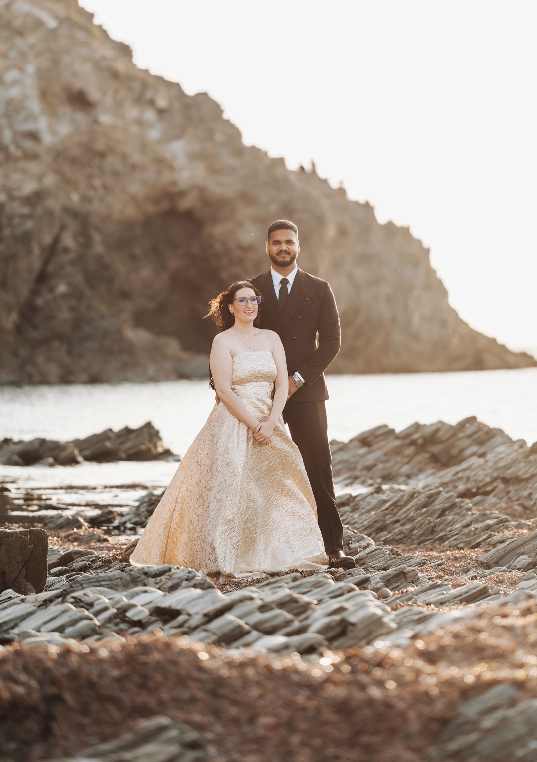 Ebony and Punit standing together on rocky shore at Second Valley, with sunlit cliffs and water behind them
