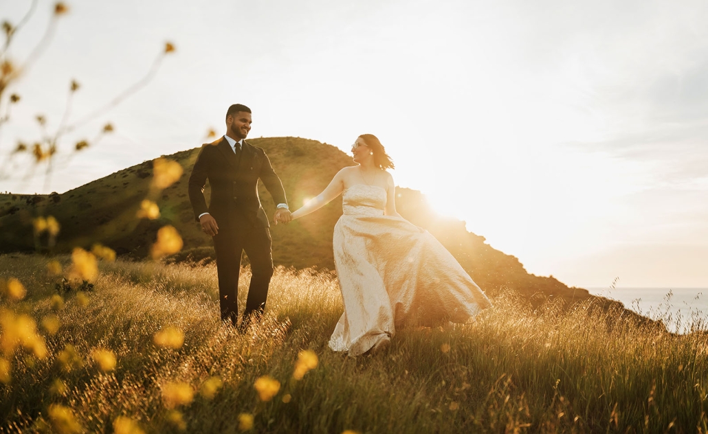 Ebony and Punit holding hands on a grassy hillside at sunset in Second Valley, with the coast behind them.