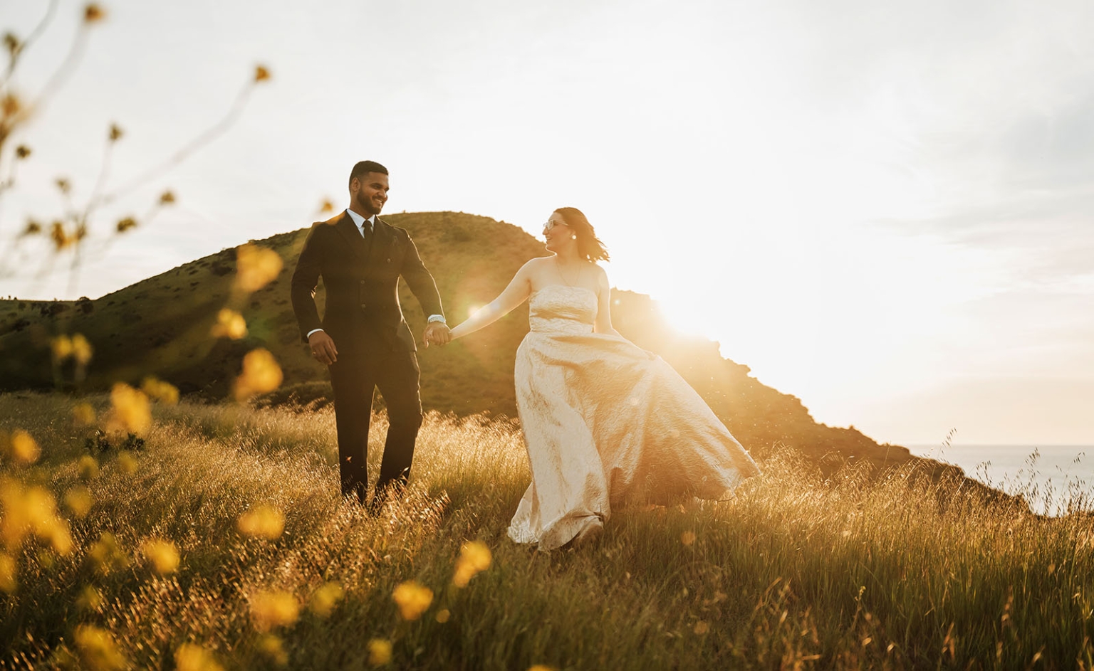 Ebony and Punit holding hands on a grassy hillside at sunset in Second Valley, with the coast behind them.