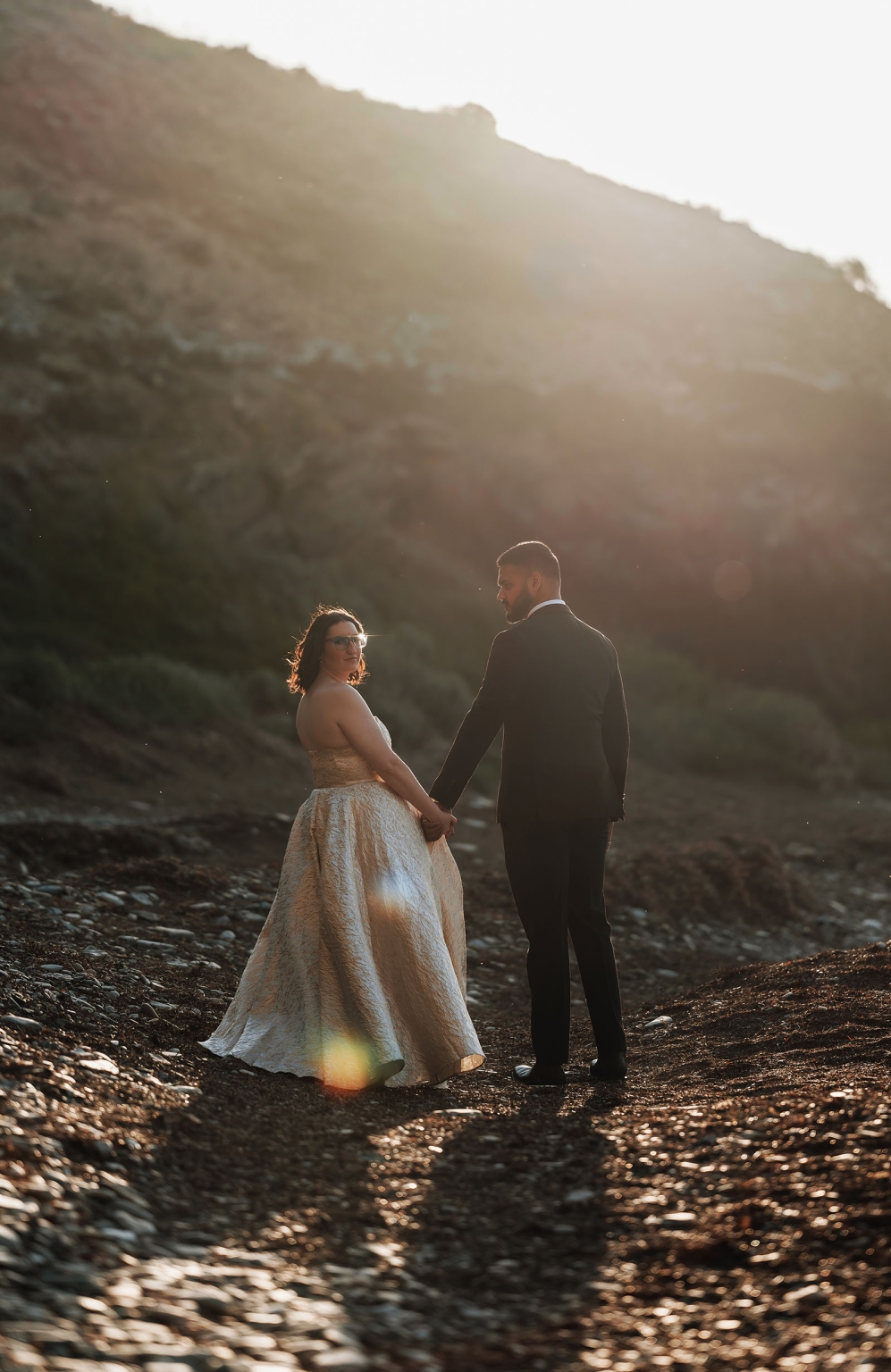 Ebony and Punit holding hands on a rocky beach beneath a sunlit hillside at Second Valley