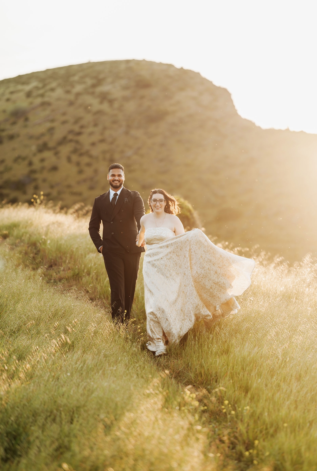 Ebony and Punit walking through sunlit grass beneath a hillside, with Ebony lifting her white wedding dress and Punit wearing a black suit.