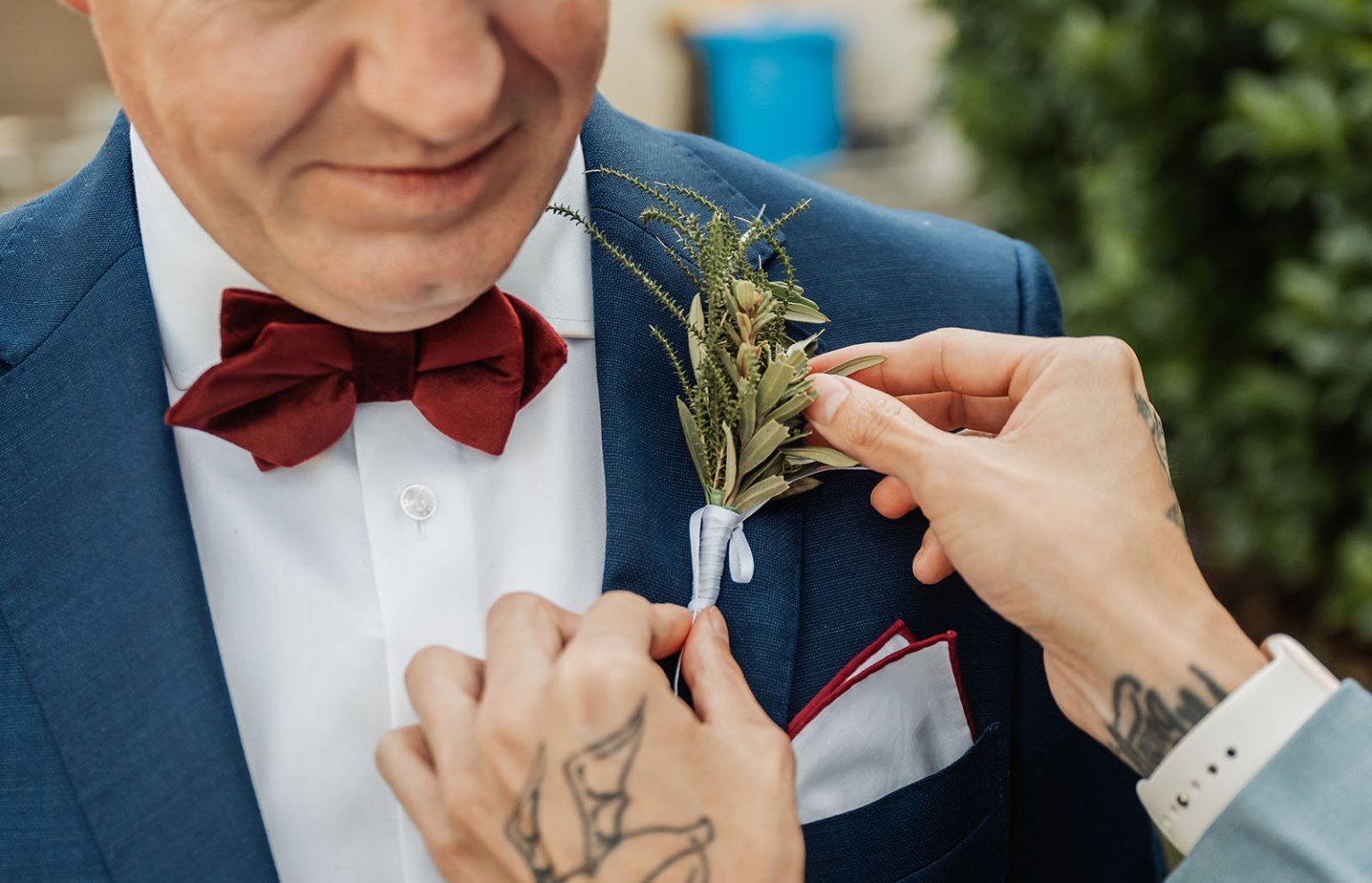hotel royal wedding - brad & manuel 3 Hands pinning a native greenery boutonniere to a groom’s blue suit jacket beside a burgundy bow tie