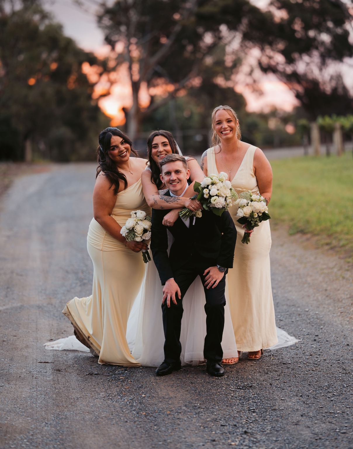 beach road winery wedding – alysha & nik 36 Groom crouching with three smiling bridesmaids holding white bouquets on a tree-lined gravel road at sunset