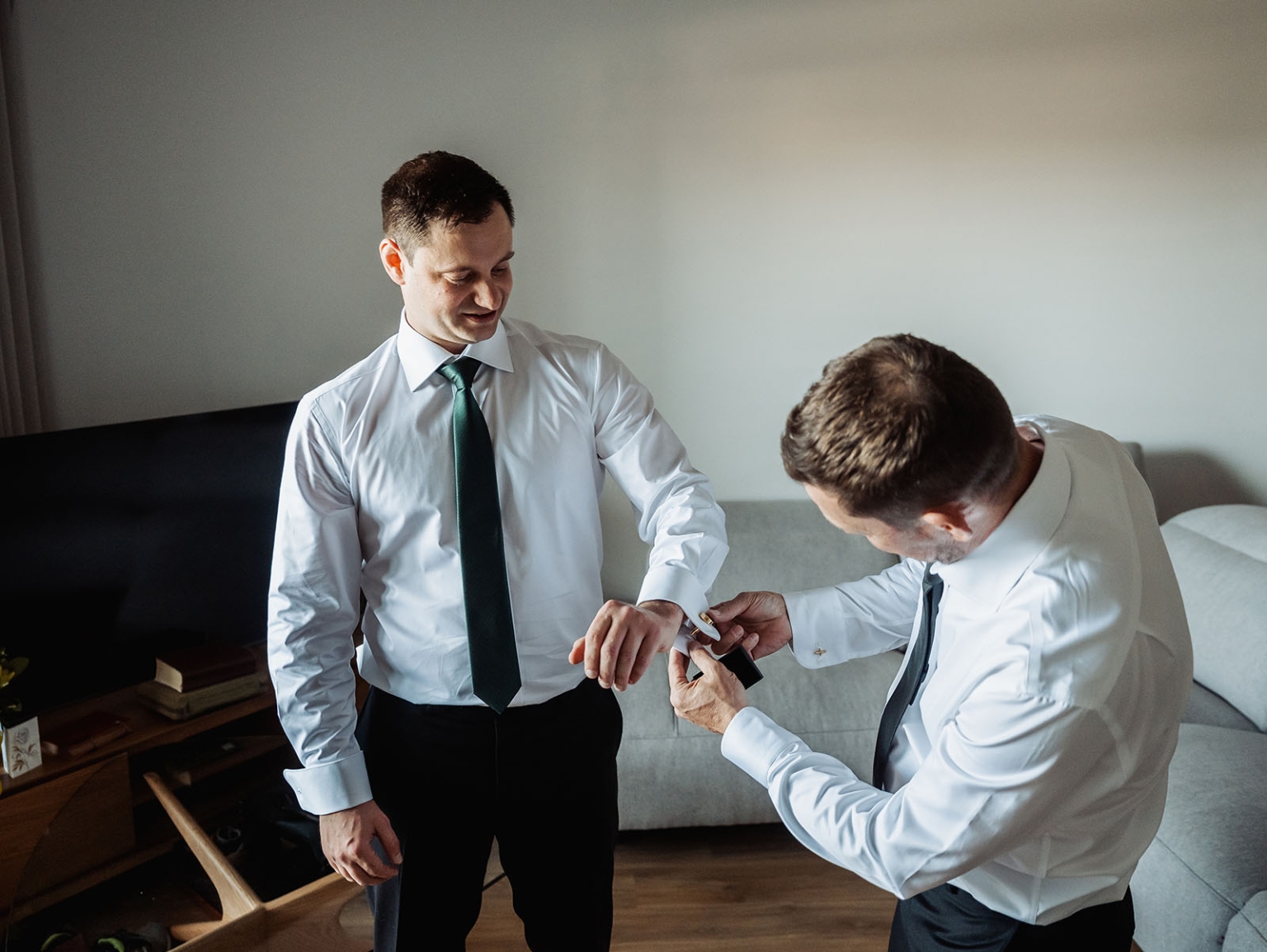 serafino winery wedding – maor & sasha 4 A man fastens cufflinks on the wrist of a groom wearing a white shirt and dark green tie in an apartment.