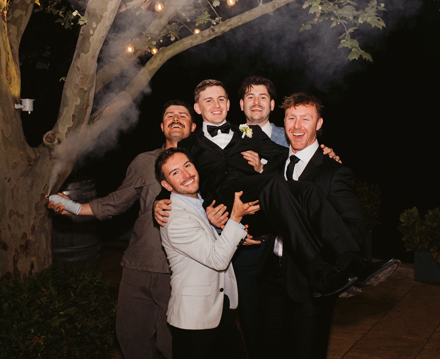 beach road winery wedding – alysha & nik 53 Wedding guests lift the smiling groom beneath a tree with festoon lights during the nighttime celebration.