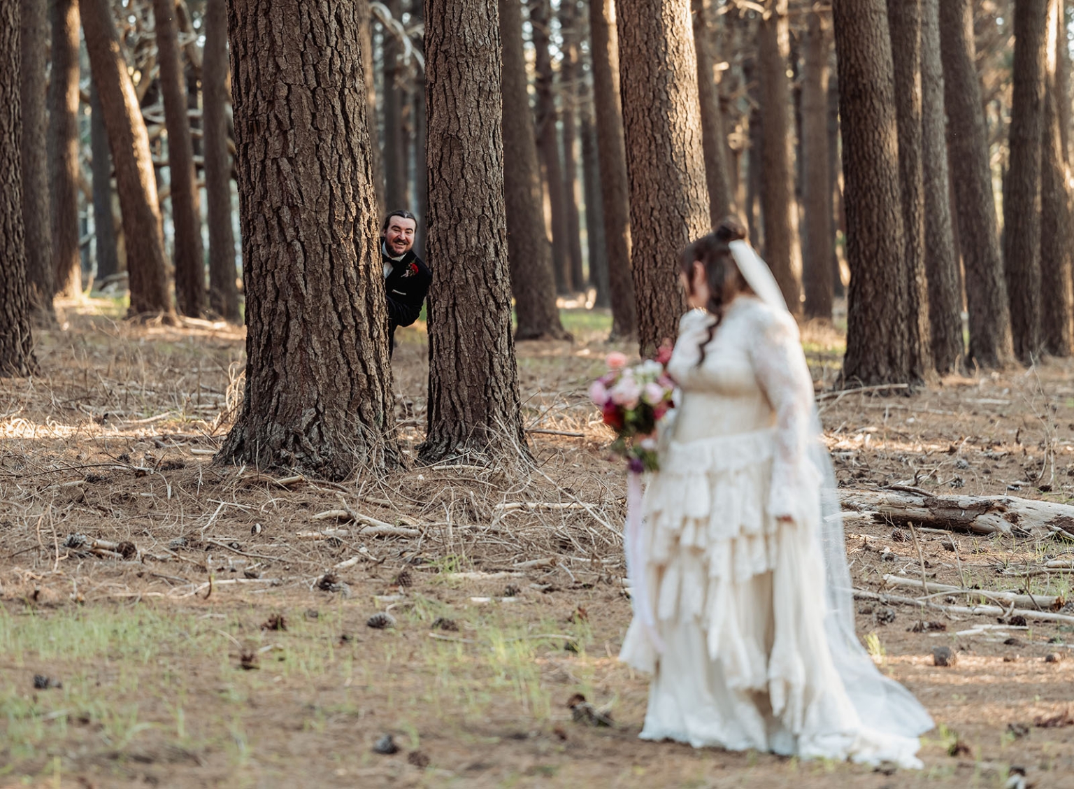 burbrook wedding – amber & angus | kuitpo forest wedding 45 Groom smiling from behind a pine tree as his bride stands in the foreground holding a colourful bouquet in Kuitpo Forest.