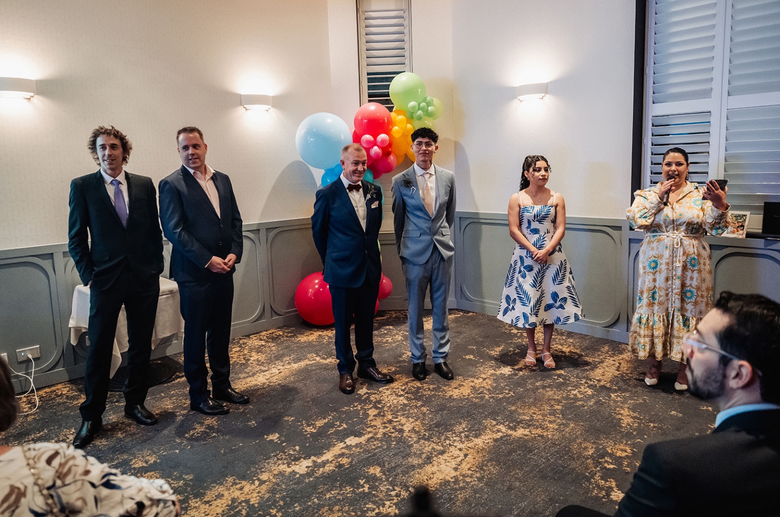 hotel royal wedding - brad & manuel 23 Wedding guests and the two grooms stand beside colourful balloons as a woman speaks into a microphone.