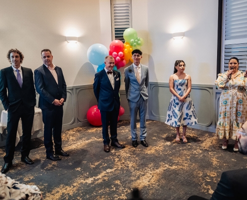Wedding guests and the two grooms stand beside colourful balloons as a woman speaks into a microphone.