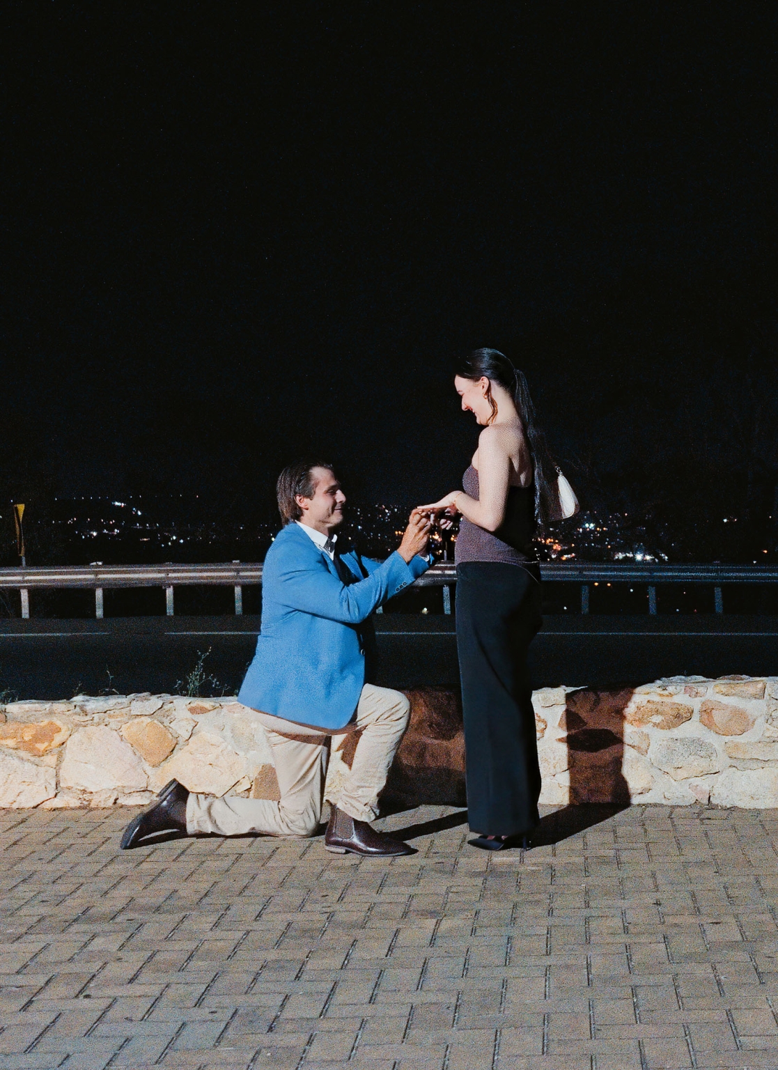 Josh kneeling and holding Kelly’s hands during a nighttime proposal at Windy Point Lookout, with Adelaide city lights behind them