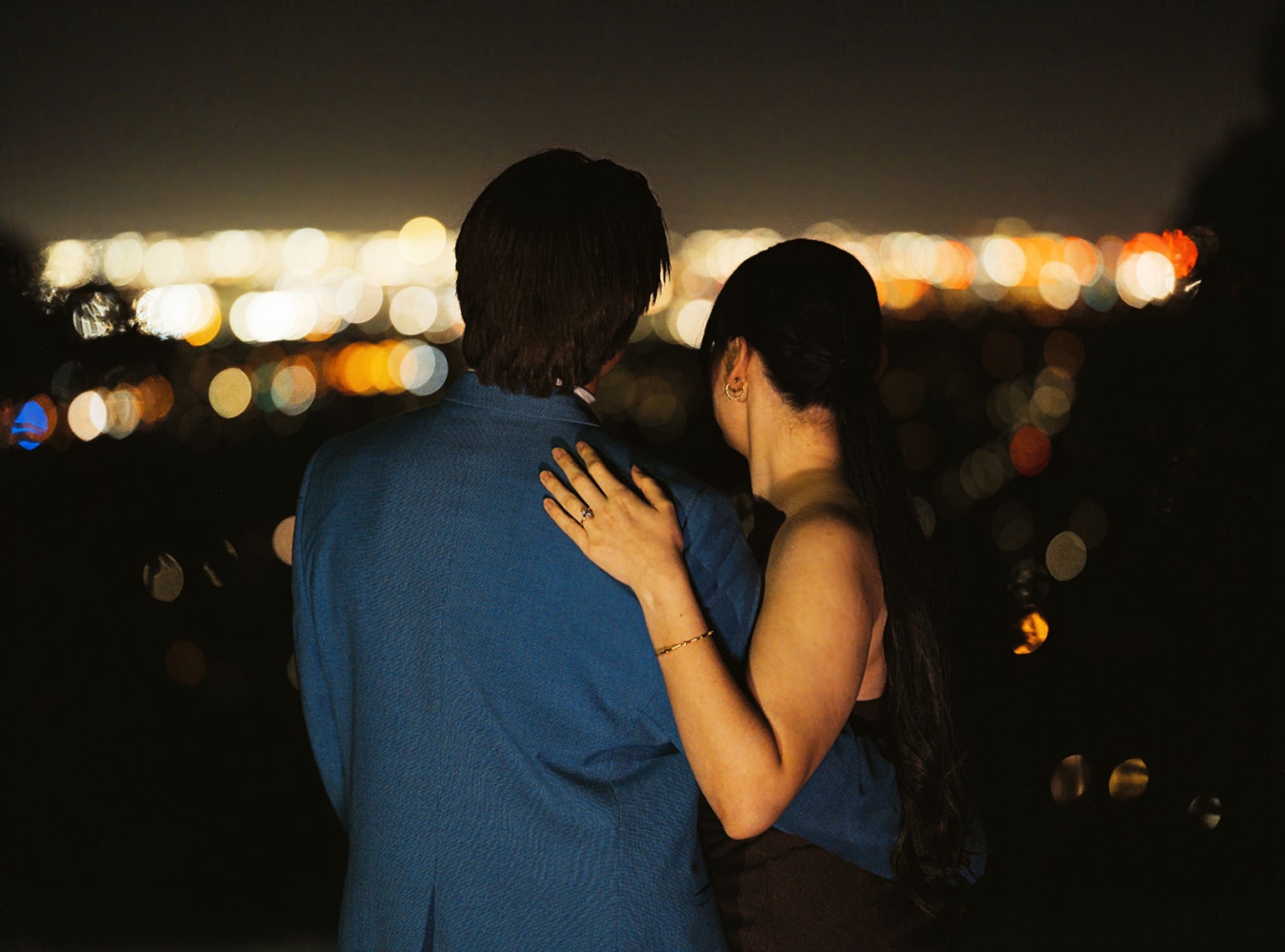 Kelly and Josh embracing at night, with her engagement ring visible against the blurred Adelaide city lights