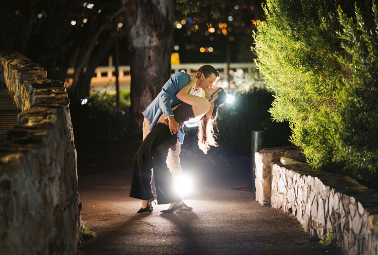 Kelly and Josh embracing on a stone-walled path at night, backlit by bright lights at Windy Point