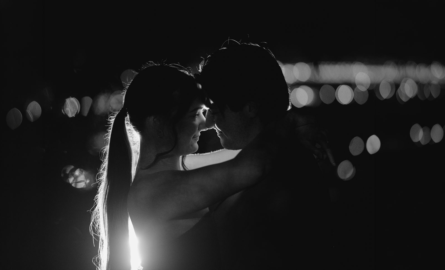 Kelly and Josh embracing forehead to forehead at night, silhouetted against Adelaide city lights at Windy Point Lookout