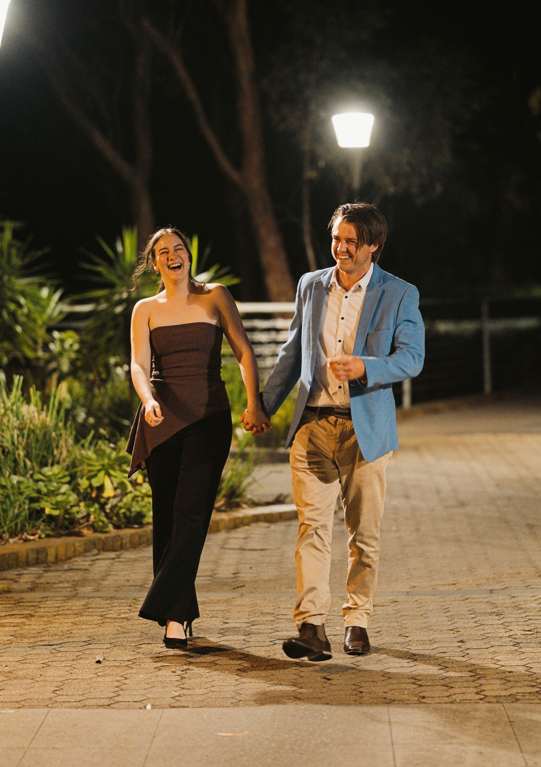Kelly and Josh laughing as they walk hand in hand along a lamp-lit garden path at night