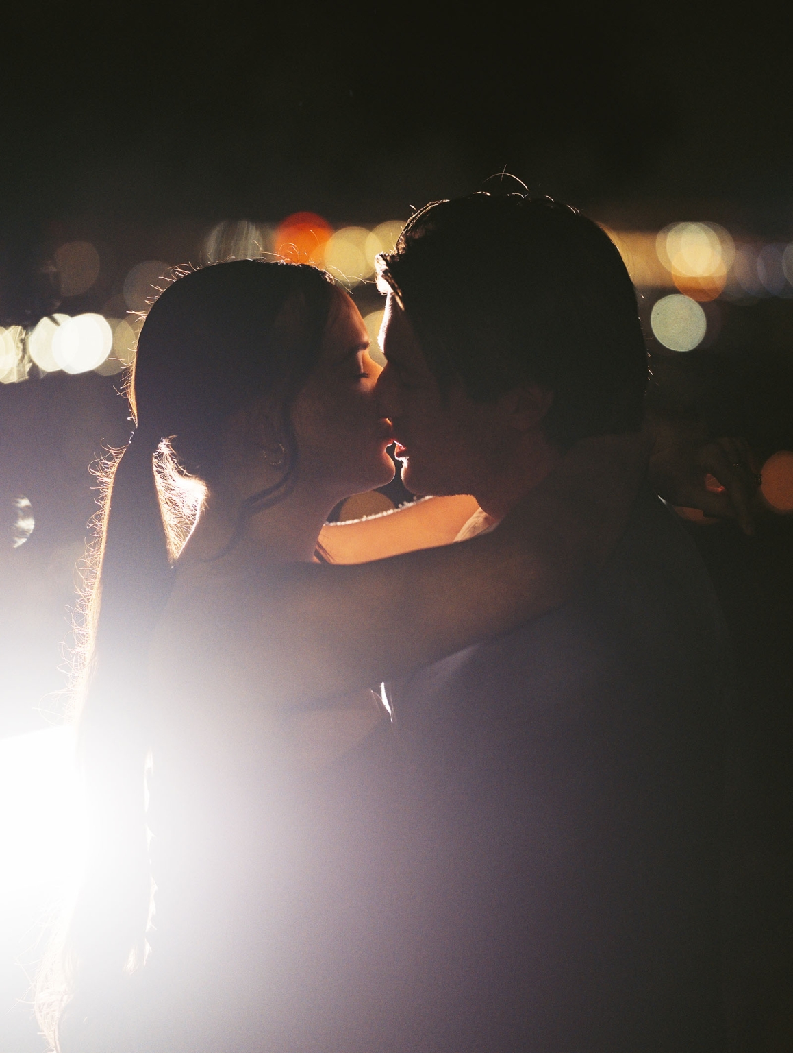 Kelly and Josh kissing at night, silhouetted against softly blurred Adelaide city lights at Windy Point Lookout.