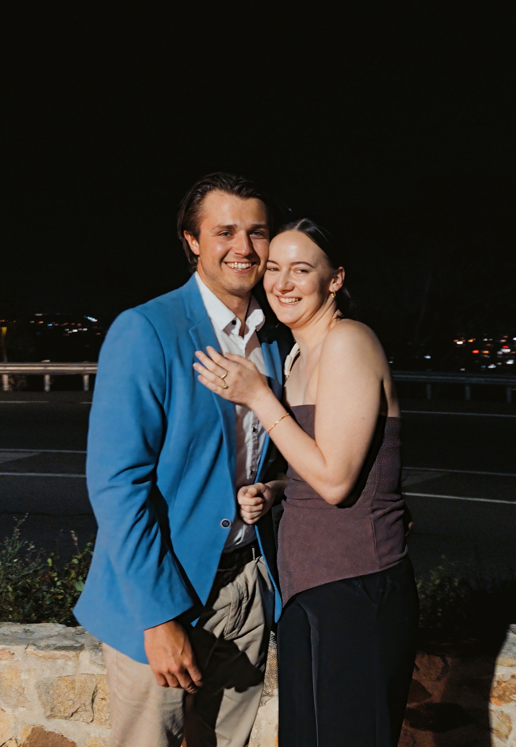 Kelly and Josh smiling together at Windy Point Lookout at night, with Kelly displaying her engagement ring and Adelaide city lights behind them.