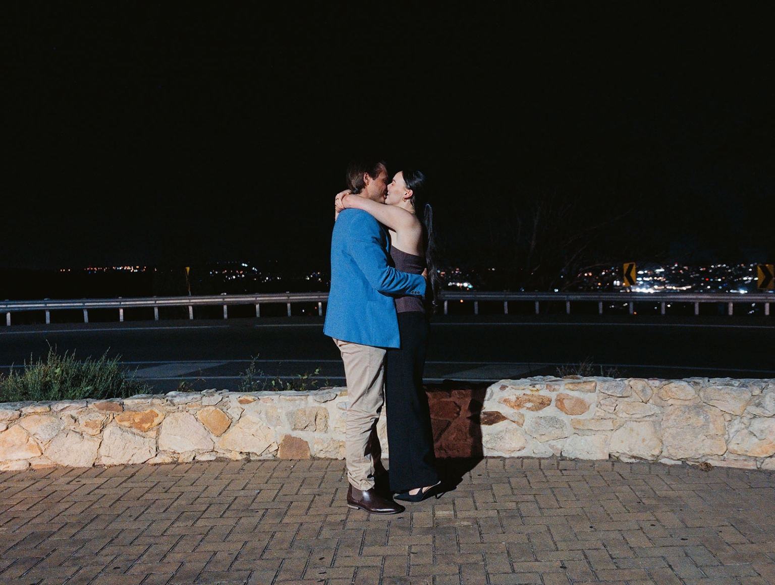 Kelly and Josh kissing beside a stone wall at Windy Point Lookout, with Adelaide city lights behind them at night