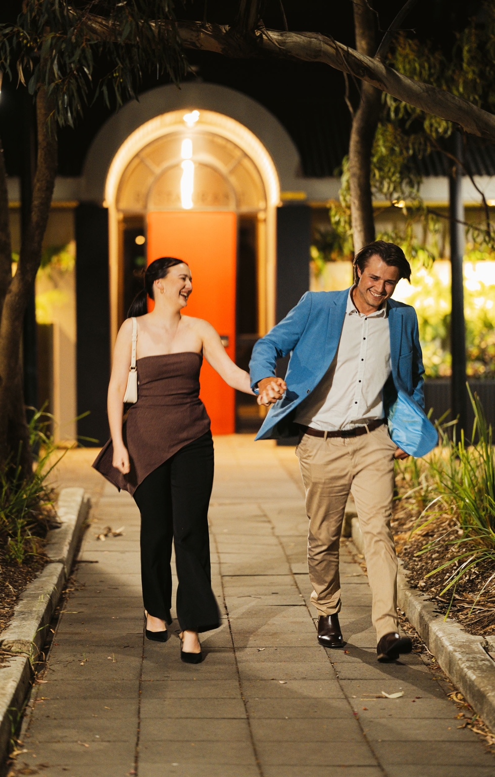 Kelly and Josh laughing as they walk hand in hand along a tree-lined path outside Windy Point Restaurant at night