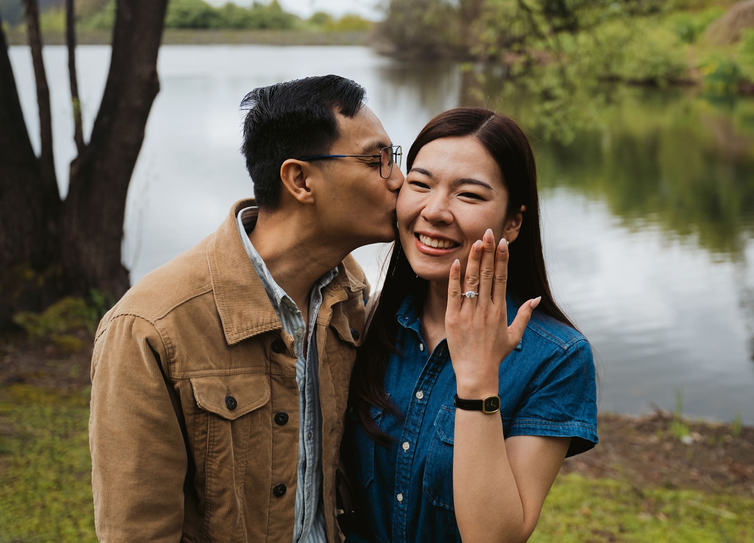 mt lofty botanic park proposal – watay & phouthun 5 Man kissing a smiling woman's cheek as she holds up her engagement ring beside a lake at Mount Lofty Botanic Garden