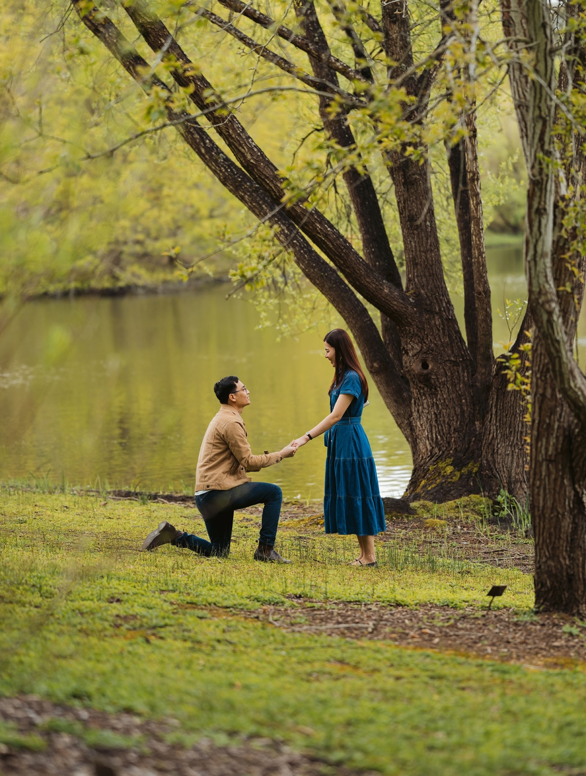 mt lofty botanic park proposal – watay & phouthun 2 Phouthun kneeling and holding Watay’s hand beside a lake beneath large trees at Mount Lofty Botanic Garden.
