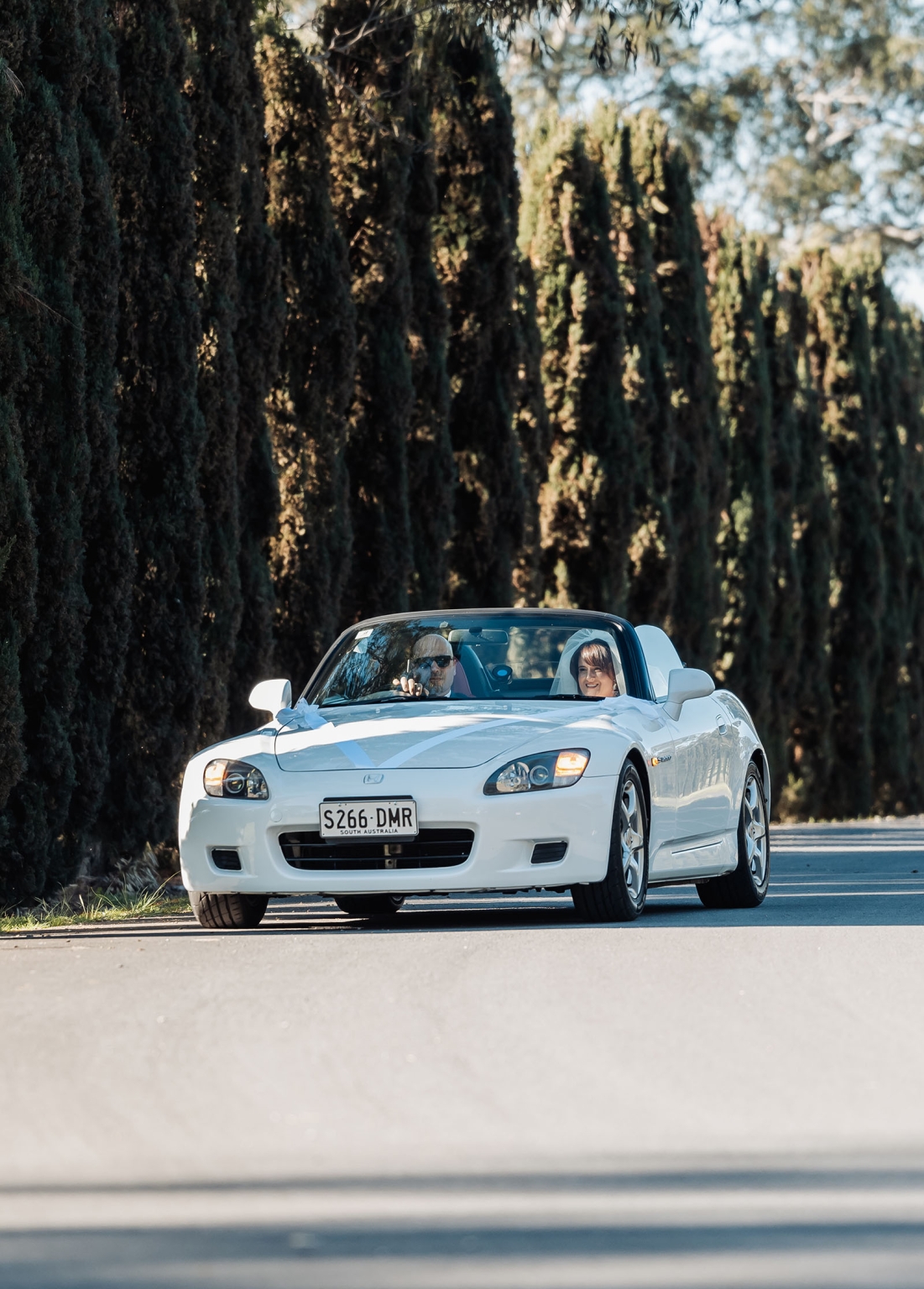 serafino winery wedding – maor & sasha 24 Newlyweds in a white convertible decorated with ribbons driving along a tree-lined road at Serafino Winery