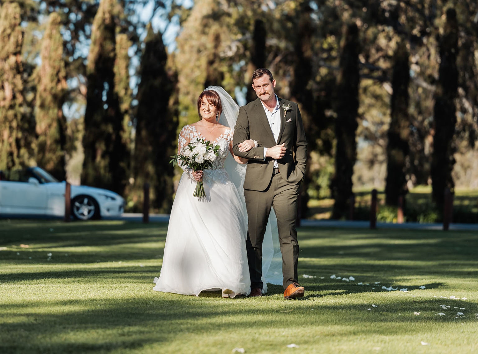 serafino winery wedding – maor & sasha 25 Bride Sasha in a lace wedding gown walks arm-in-arm with a man across the lawn at Serafino Winery, holding a white bouquet.